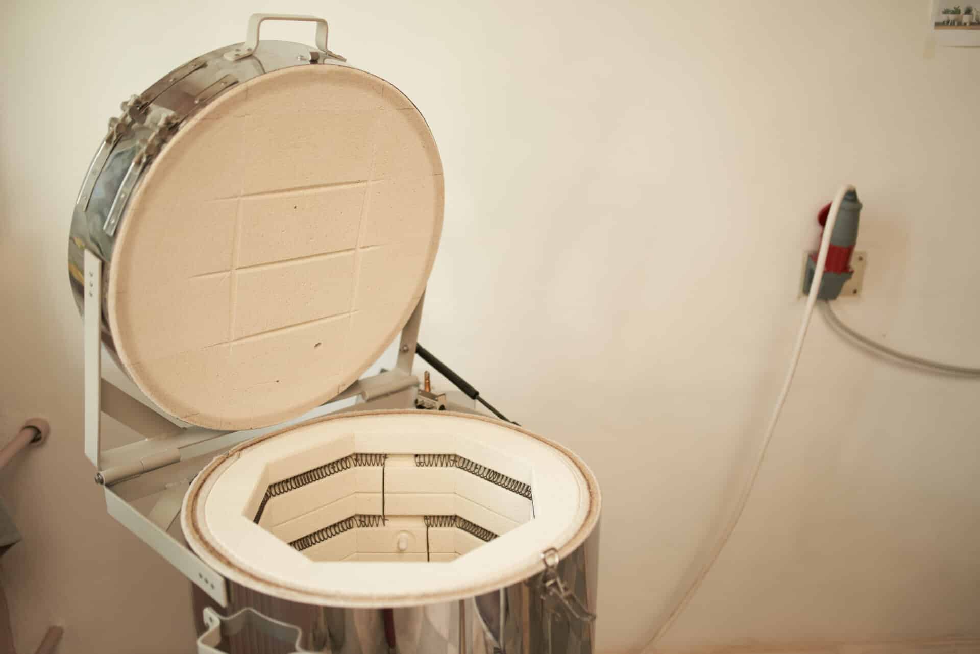 Modern electric kiln with opened lid located near wall and connected with socket in workshop